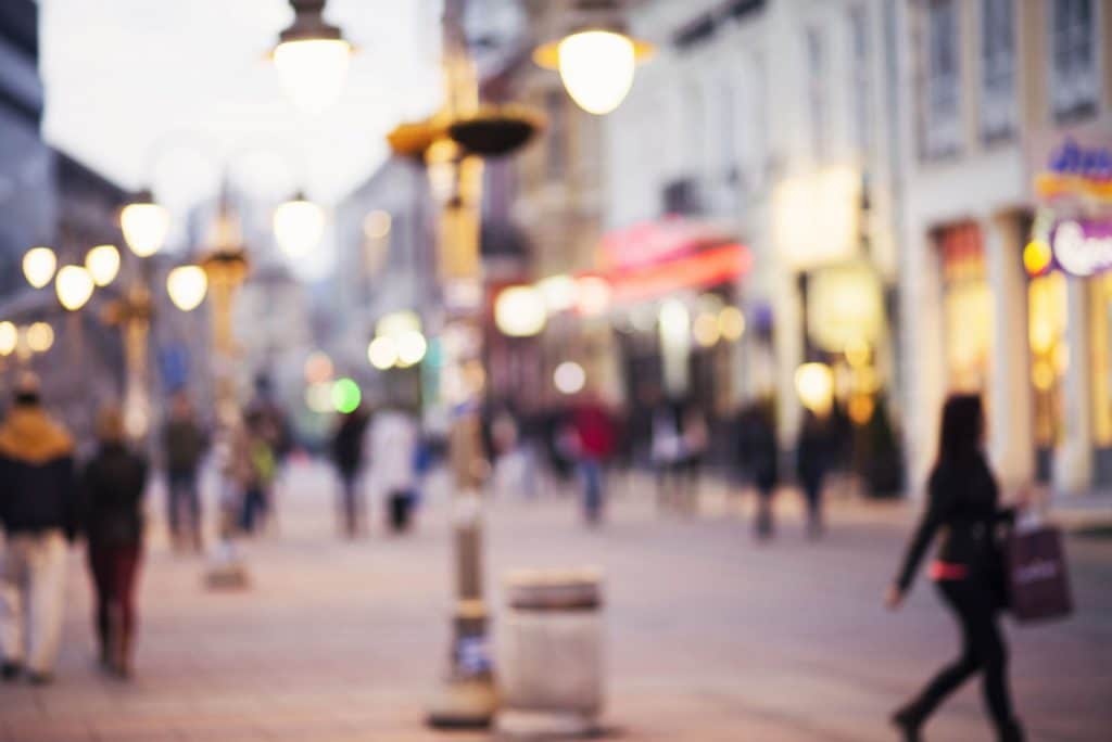 Foto de uma rua de uma cidade grande, com algumas pessoas passando com sacolas nas mãos e os postes de luz acesos.