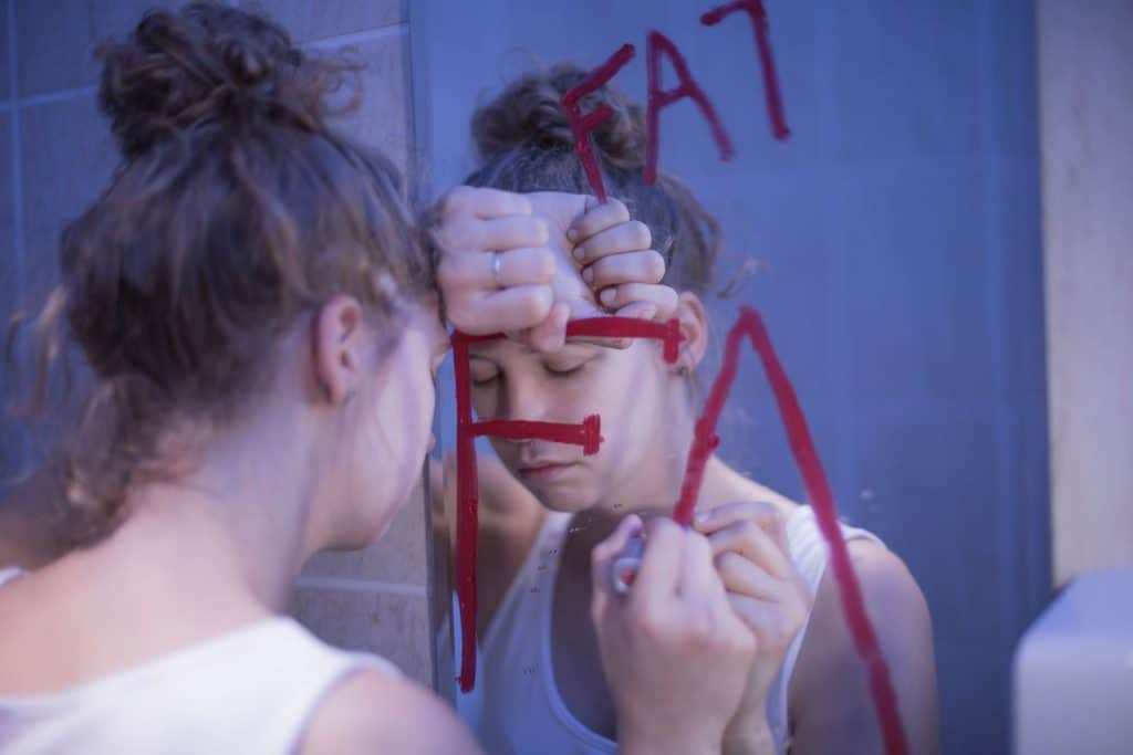 Mulher branca com os cabelos presos em um coque apoiando a cabeça sobre o braço em frente a um espelho. A mulher está com uma expressão triste e segura um batom vermelho em sua mão direita que foi utilizado para escrever a palavra "fat" no espelho, que significa "gordo" em inglês.