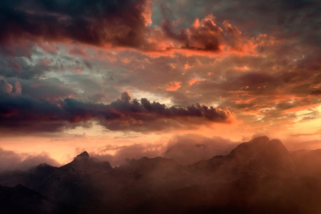 Uma paisagem com foco no céu que está alaranjado, porque é um final de tarde, e há muitas nuvens. Abaixo do céu é possível ver a silhueta de algumas montanhas.