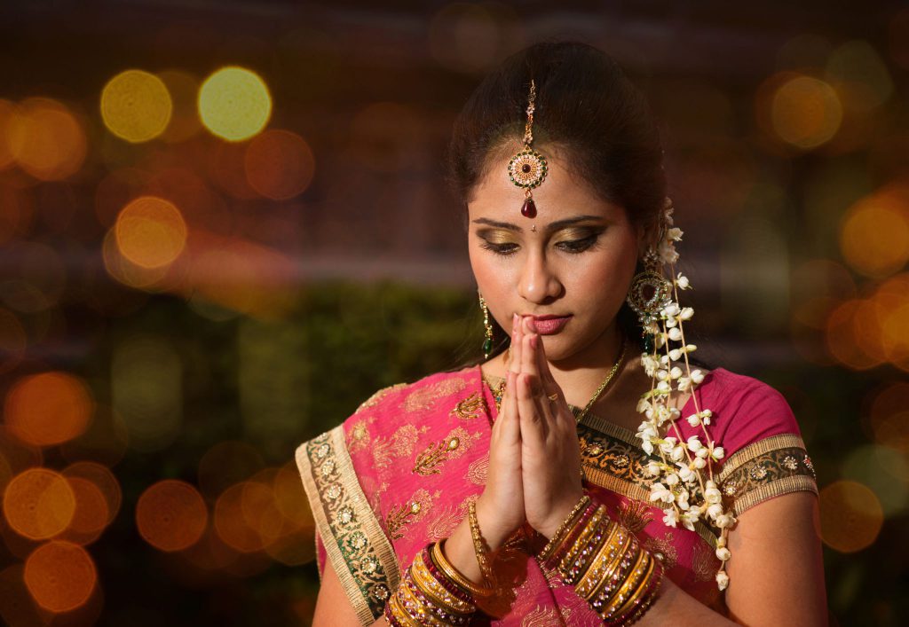 Mulher com vestes e maquiagens indianas fazendo o sinal de namastê com as mãos.