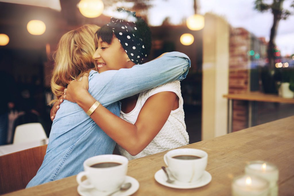 Duas mulheres em um café se abraçando. Uma delas é loira e está vestindo uma camisa de manga longa azul clara e a outra é morena e está vestindo uma camiseta de manga curta branca, está com uma bandana preta com bolinhas brancas na cabeça e também está usando um bracelete dourado. Há também duas xícaras de café em cima de um balcão que está ao lado das duas moças.