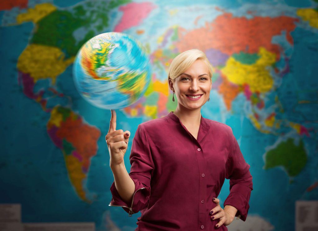 Mulher branca e loira com o cabeço preso em um coque e ela está usando uma camisa social cor de vinho. Ela está com uma mão na cintura e a outra está com o dedo indicador levantado equilibrando um globo terrestre e ao fundo há um mapa do mundo.