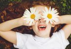 Menina de cabelos ruivos e encaracolados deita no chão. Ela está sorrindo e está segurando uma flor margarida em cada mão e as coloca na altura do olho.