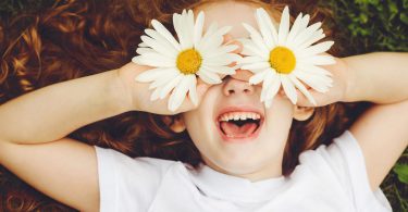 Menina de cabelos ruivos e encaracolados deita no chão. Ela está sorrindo e está segurando uma flor margarida em cada mão e as coloca na altura do olho.
