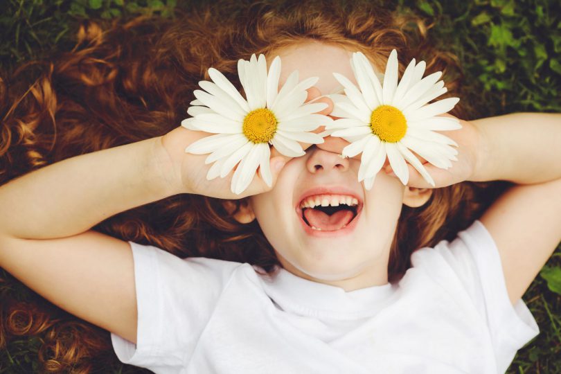 Menina de cabelos ruivos e encaracolados deita no chão. Ela está sorrindo e está segurando uma flor margarida em cada mão e as coloca na altura do olho.