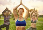 Três mulheres em posição de Yoga, com as mãos se tocando acima da cabeça. Há uma mulher no centro que é loira e está vestindo um top azul, a sua direita há uma mulher negra vestindo uma blusa de manda comprida preta com listras rosas, vermelhas e brancas. A esquerda há outra mulher loira vestindo um top verde musgo. Ao fundo há um fim de tarde, árvores e gramado.