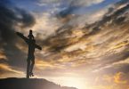 Imagem de um crucifixo em cima de um pequeno monte com um céu nublado ao fundo.