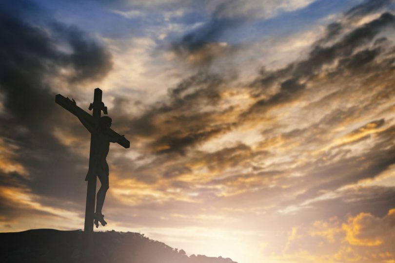 Imagem de um crucifixo em cima de um pequeno monte com um céu nublado ao fundo.