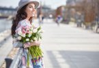 Mulher de olhos fechados, sorrindo, segurando um buquê de flores, enquanto atravessa a rua.