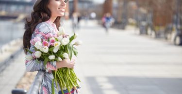 Mulher de olhos fechados, sorrindo, segurando um buquê de flores, enquanto atravessa a rua.