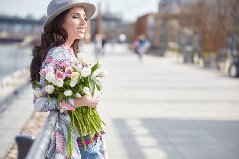 Mulher de olhos fechados, sorrindo, segurando um buquê de flores, enquanto atravessa a rua.