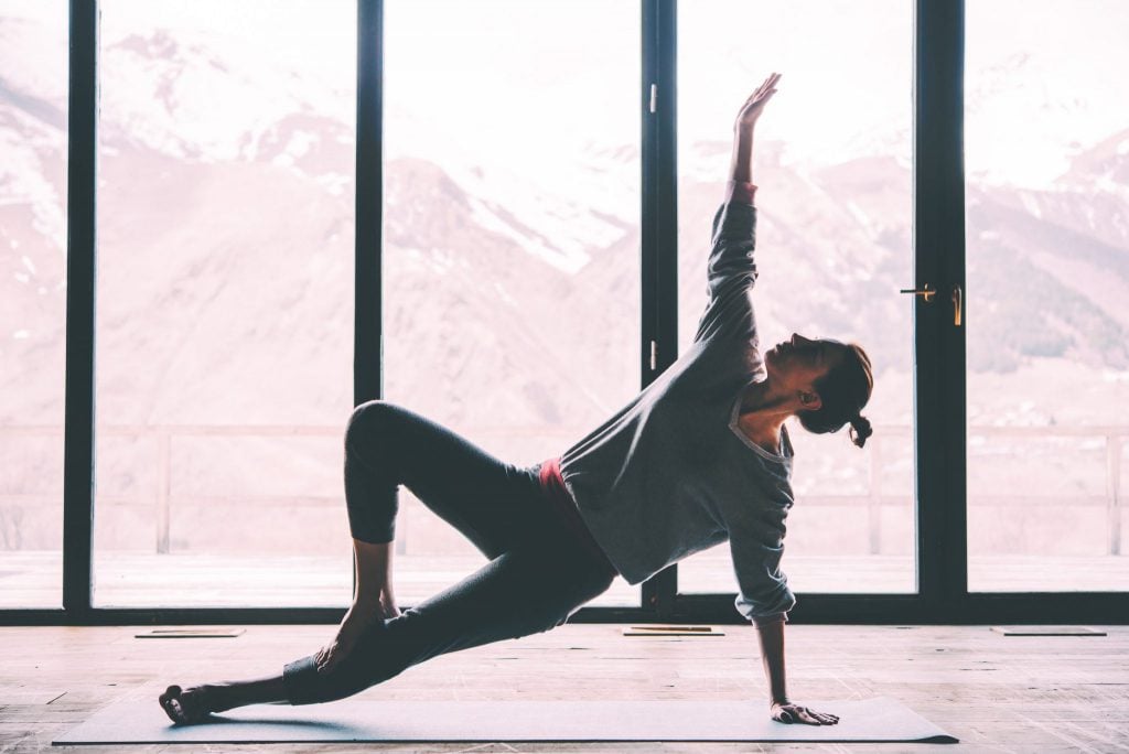 Mulher de cabelos escuros presos em um coque, que está dentro de uma sala com grandes janelas e está em cima de um tapete de yoga. A mulher está apoiada em um braço de forma lateral enquanto o outro está esticado para cima, uma perna está esticada dando apoio enquanto a outra está semi-dobrada acima da que está esticada.