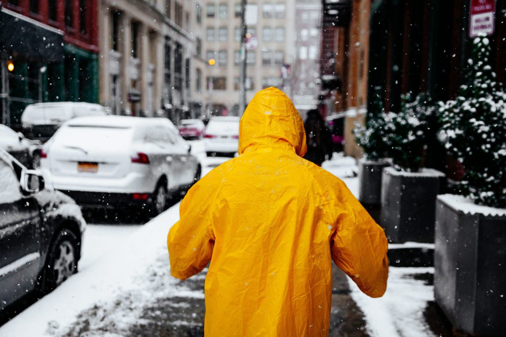 Homem de costas vestindo uma capa de chuva amarela. Ao fundo o cenário é de uma rua com carros estacionados e cobertos pela neve.