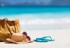 Bolsa, chapéu de palha, óculos de sol e chinelo azul em areia da praia, com mar ao fundo.