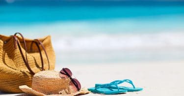 Bolsa, chapéu de palha, óculos de sol e chinelo azul em areia da praia, com mar ao fundo.