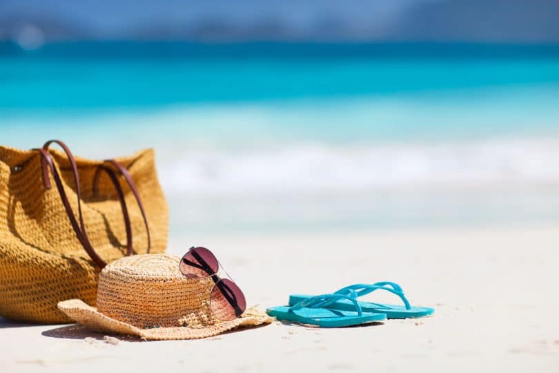 Bolsa, chapéu de palha, óculos de sol e chinelo azul em areia da praia, com mar ao fundo.