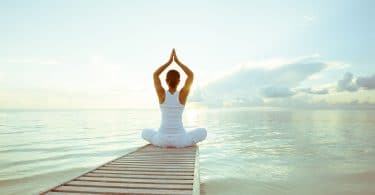 Mulher branca sentada em uma posição de yoga em cima de um pier em frente ao mar.