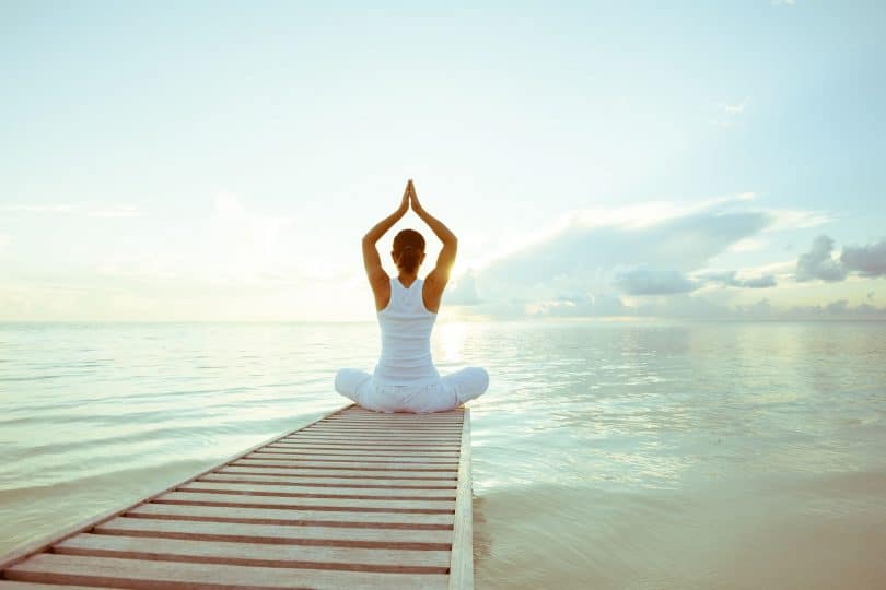 Mulher branca sentada em uma posição de yoga em cima de um pier em frente ao mar.