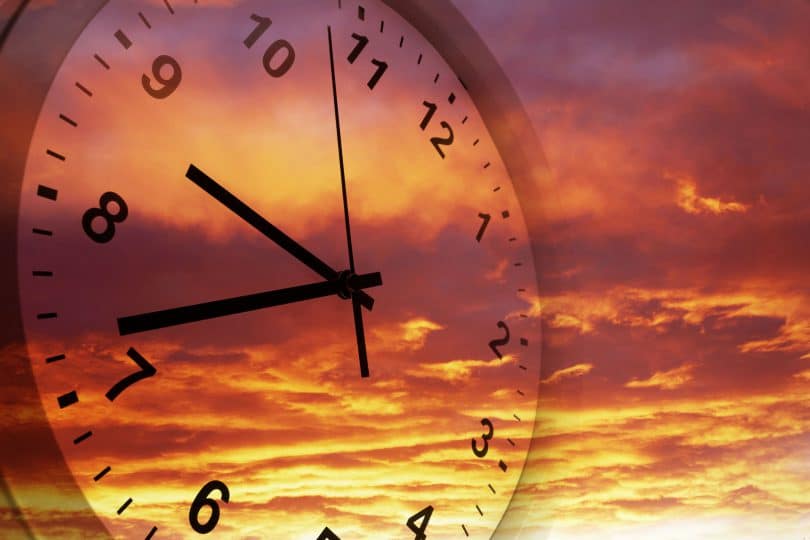 Imagem de relógio de parede com um pôr do sol avermelhado ao fundo.