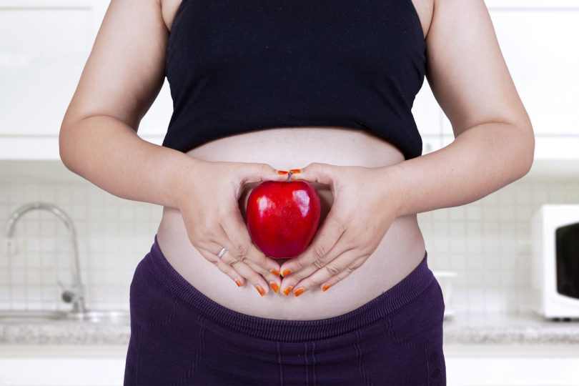 Mulher grávida segurando uma maça vermelha em frente da sua barriga.