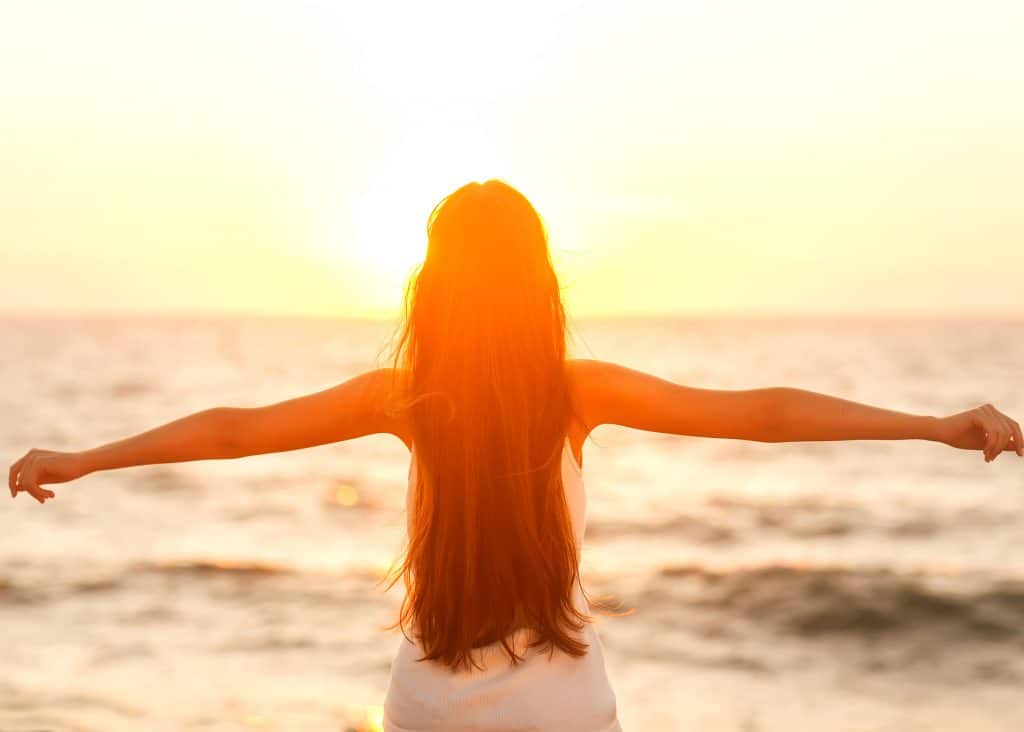 Mulher de costas com os braços abertos e ao fundo há a paisagem de uma praia ao fim de tarde.