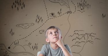 Menino branco e loiro, sentado em uma mesa, com um livro aberto, com vários desenhos de mapa ao seu fundo.