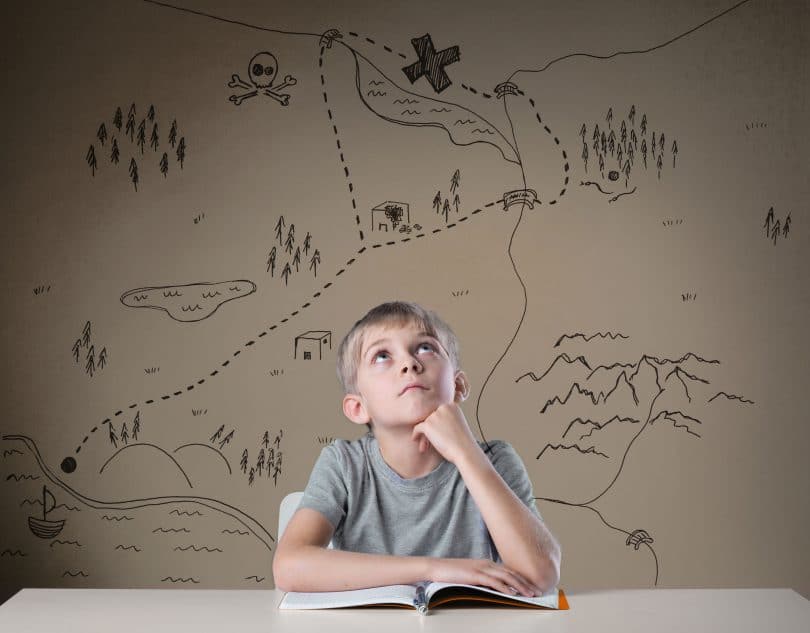 Menino branco e loiro, sentado em uma mesa, com um livro aberto, com vários desenhos de mapa ao seu fundo.