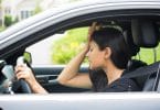 Mulher dentro do carro, vestida de preto , com a face preocupada e com a mão na cabeça.