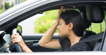 Mulher dentro do carro, vestida de preto , com a face preocupada e com a mão na cabeça.