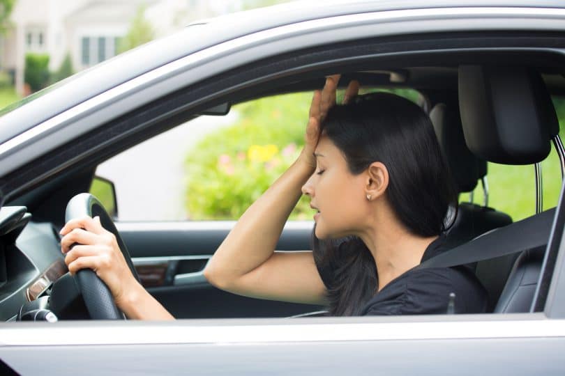 Mulher dentro do carro, vestida de preto , com a face preocupada e com a mão na cabeça.