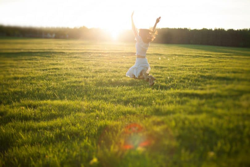 Mulher com os cabelos soltos, correndo em um campo verde, com os braços pra cima e luz do sol brilhando ao fundo.