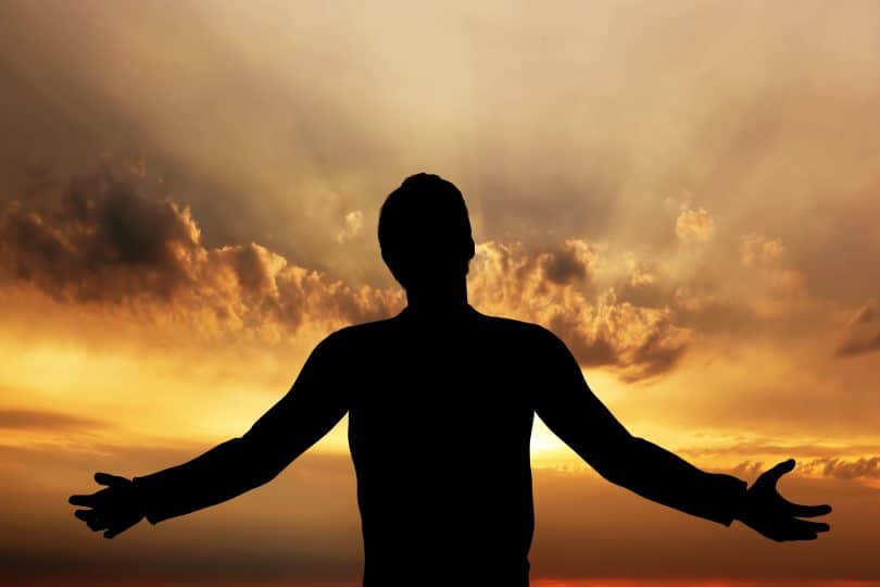 Silhueta de homem com os braços abertos em frente à um céu iluminado pelo pôr do sol.