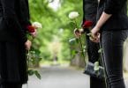 Pessoas vestidas de preto, em um cemitério, segurando rosas vermelhas e brancas.