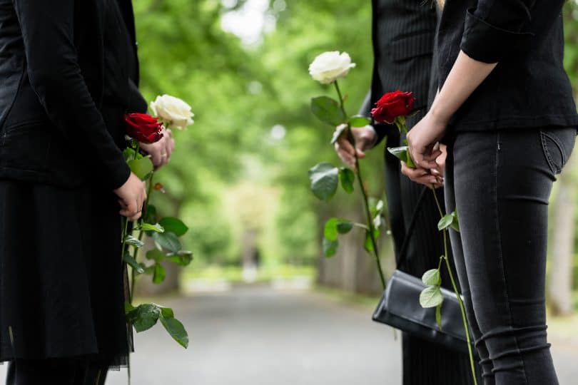 Pessoas vestidas de preto, em um cemitério, segurando rosas vermelhas e brancas.