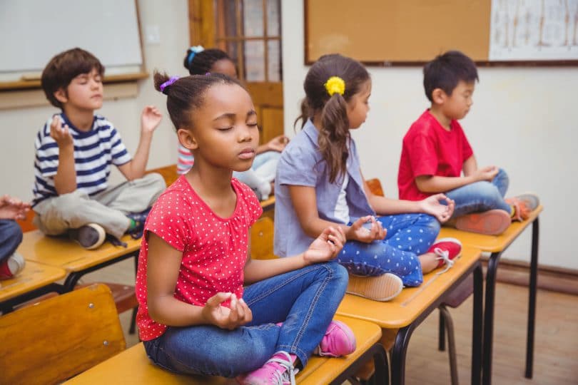 Crianças em posição de meditação em cima de suas mesas escolares.