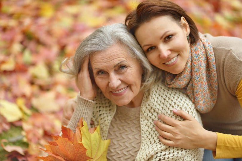 Mãe idosa e filha abraçadas, sorrindo, com folhas de outono ao fundo.