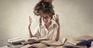 Mulher vestida de branco, com os cabelos em pé, gritando com as mãos pra cima, cercada de livros abertos .