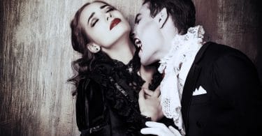 Homem vestido de vampiro, com roupas de época, mordendo o pescoço de uma mulher jovem, vestida com roupas de época.