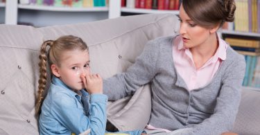 Criança menina sentada em um sofá, triste com a mão na boca enquanto a sua mãe olha pra ela e coloca a mão no seu ombro.