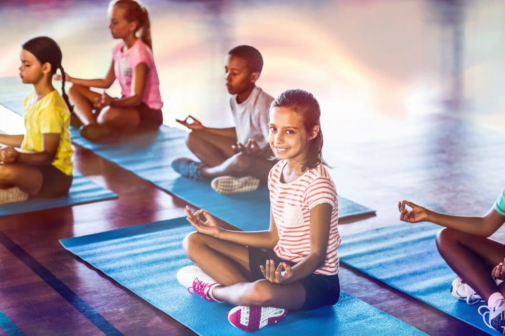 Crianças sentadas em tapetinhos de yoga azuis claro e em posição de meditação.