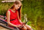 Menina vestida de vermelho, sentada no chão, em cima de uma bicicleta, enquanto mexe em um tablet, em um parque, com um lago ao fundo.