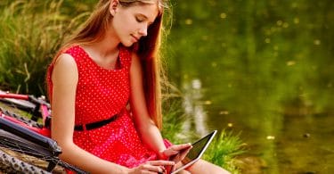Menina vestida de vermelho, sentada no chão, em cima de uma bicicleta, enquanto mexe em um tablet, em um parque, com um lago ao fundo.
