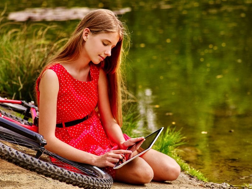 Menina vestida de vermelho, sentada no chão, em cima de uma bicicleta, enquanto mexe em um tablet, em um parque, com um lago ao fundo.
