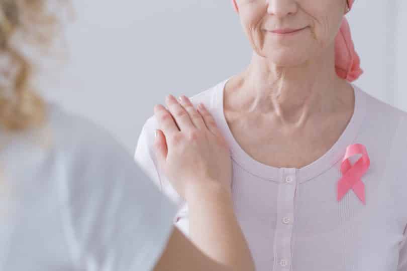 Mulher apoiando o braço sobre o ombro de uma mulher mais velha, com lenço na cabeça, que sofre de câncer de mama.