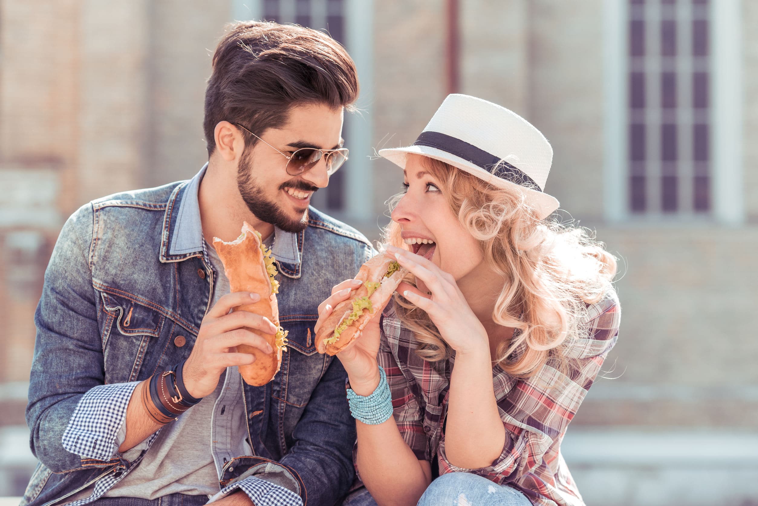 Casal, de homem e mulher, sentados no chã de uma praça, olhando um para o outro, rindo e comendo sanduíches. sanduíche