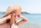 Mulher passa protetor solar no rosto. Ela está de olhos fechados, sorri e veste um chapéu. O ambiente é de praia.