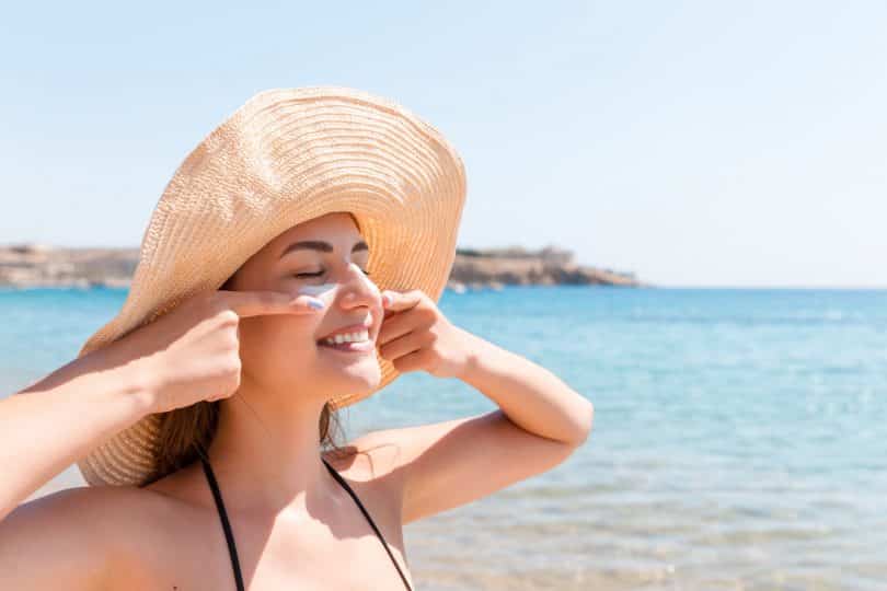 Mulher passa protetor solar no rosto. Ela está de olhos fechados, sorri e veste um chapéu. O ambiente é de praia.