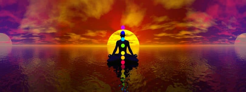 Desenho gráfico de um homem em meditação. Chakras representados como bolas coloridas. Fundo de pôr-do-sol e mar.