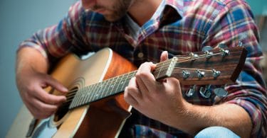 Close em um músico tocando um violão. Ele veste uma blusa xadrez.