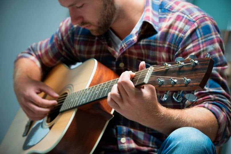 Close em um músico tocando um violão. Ele veste uma blusa xadrez.